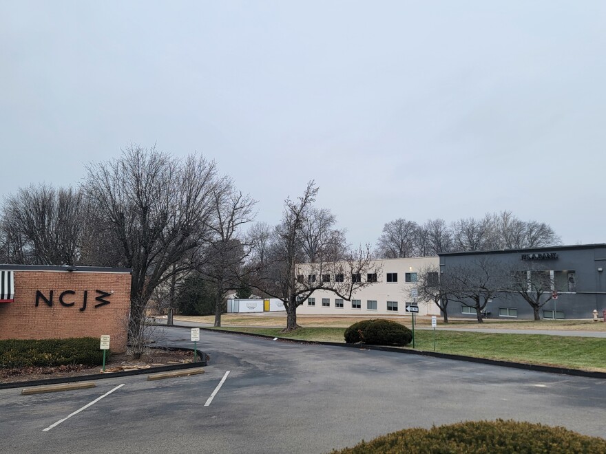 A light brick building with black lettering can  be seen in the photo to the left. A two-story building that is gray in the front and white in the back is in the background. A parking lot and side street separate the two buildings.