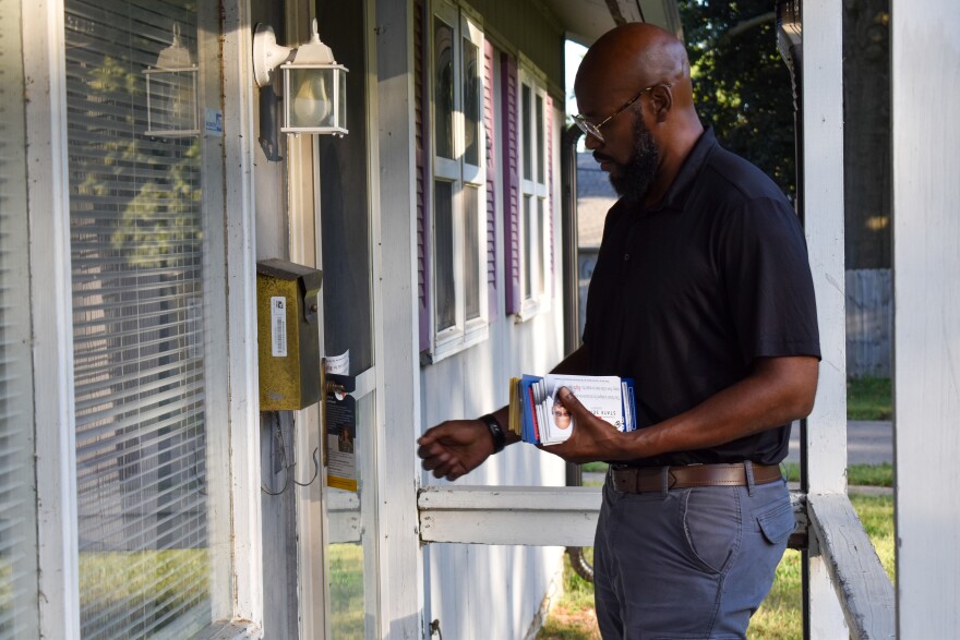 A black man in a black polo and grey slacks knocks on the front door of a suburban house.