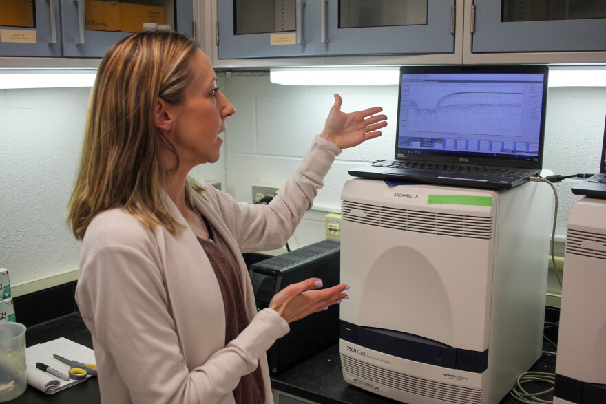 A woman in a laboratory gestures to a computer screen that displays line graphs. 