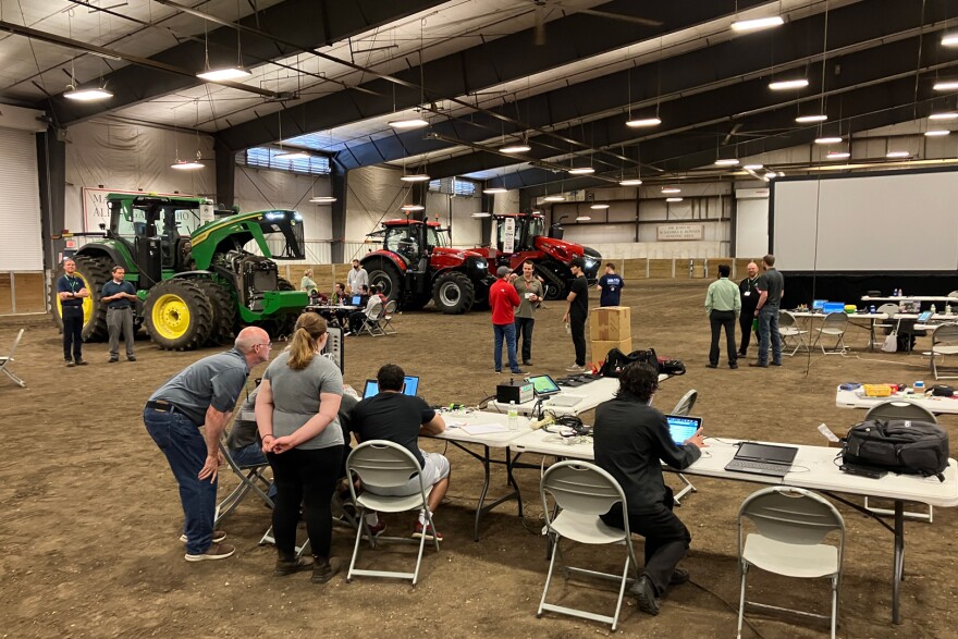 People sit at folding tables to work on laptops in an arena with dirt. Tractors are lined up in the background.