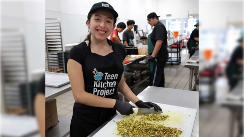 A smiling young woman with a baseball cap and apron wears black food-service gloves as she chops a pile of something on a white cutting board. Behind her more young adults work in a commercial kitchen filled with stainless steel counters and pans. 