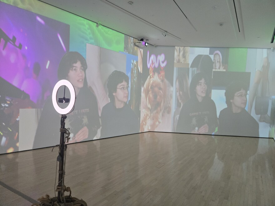 gallery space with a selfie camera and ring light in the center, on the walls are projected images of museum patrons and social media videos