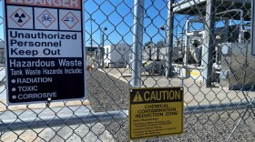 A silver metal fence is pictured, with various warning signs about hazardous waste and industrial structures in the background 