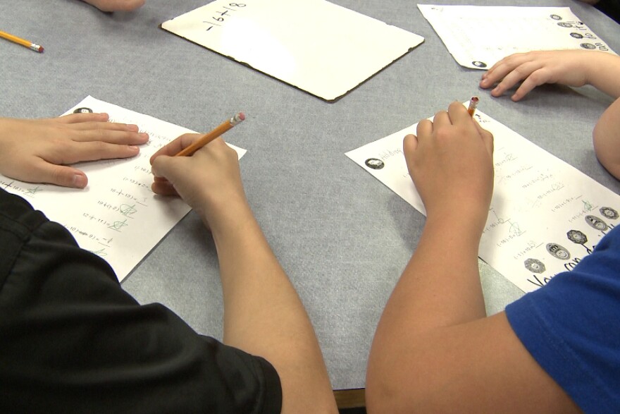 Children complete worksheets at a table in a classroom.