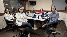 Five people wearing headphones sit at a table in a radio talk studio: a woman at foreground left has long dark hair and is wearing glasses and a white button-down shirt; a woman at foreground right has short dark hair and is wearing a beige cardigan sweater, white shirt, jeans and brown flats; a man at center has short dark hair and is wearing a grey button-down shirt, jeans and brown shoes; a man at background left has short brown hair and is wearing glasses and a brown button-down shirt; a woman at background right has long blonde hair and is wearing glasses, a red jacket and a black and white striped shirt.