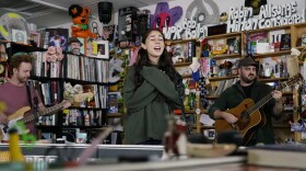 A young woman crosses her arms over her chest and sings with her eyes closed while two men play guitars on either side of her in the background. All three are behind an office desk.