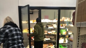 Several people shop for food at a food bank. A woman sorts through goods in a shopping cart, and a man stands in front of a food cooler, holding the door open. In the foreground is a shopping cart full of goods.