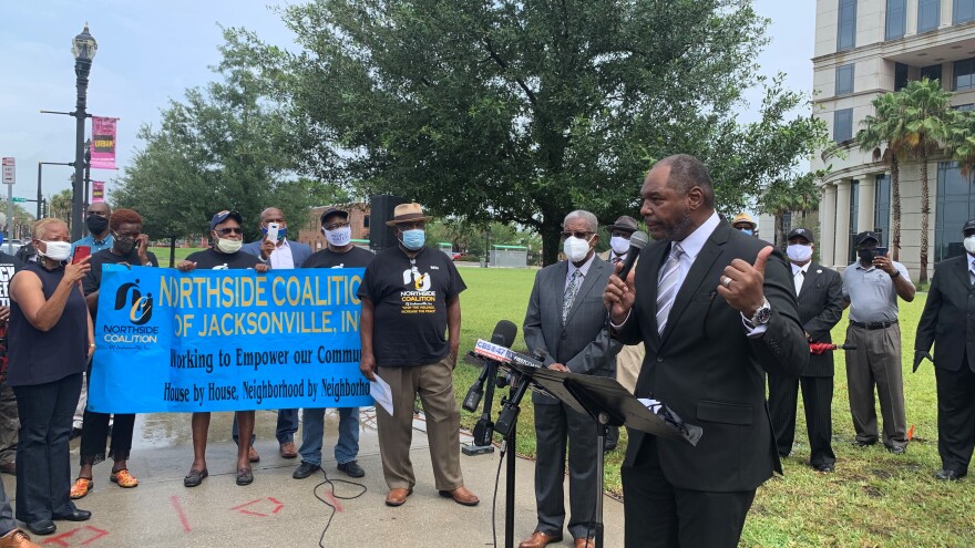 John Allen Newman talking into the camera, pointing behind him. Off to the left is a sign for the Northside Coalition of Jacksonville. Other people standing around Newman