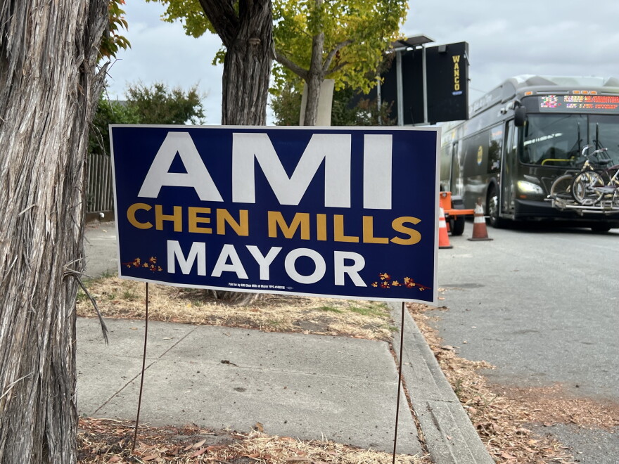 A purple and white sign is stuck in a patch of dirt next to a tree trunk. A bus drives on the street in the right corner.