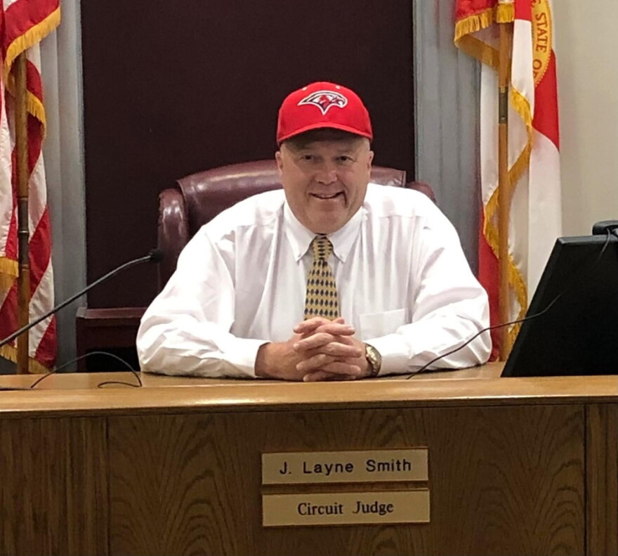 Judge Layne Smith wears a ball cap and sits in his courtroom, smiling for the camera