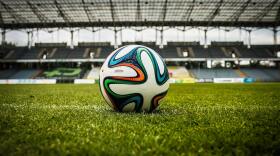 A close-up view of a soccer ball resting on a green field inside an open-air stadium, with empty stands and a steel canopy visible in the background.