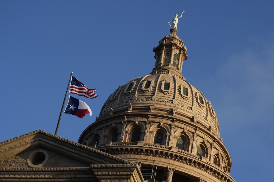  A close-up photo of the tan stone dome of the Texas Capitol building. A statue of a woman holding a five-pointed star stands on top of the dome. Just in front of the dome, an American flag flies above a Texas flag.