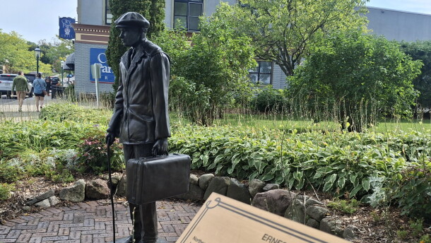 A statue of a young man with a suitcase and cane is visible in a park in downtown Petoskey. In the foreground, a gold plaque reads "Ernest Miller Hemingway." 