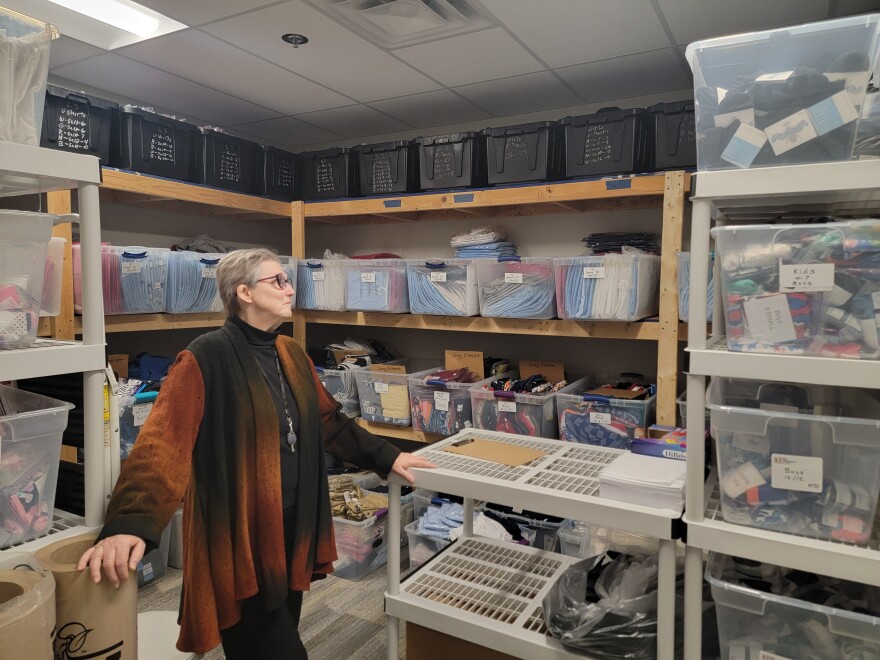 An older white woman in black pants a black shirt and a brown and black sweater stands in a storage room. Clear and black bins on wood and plastic shelves hold school uniforms, socks, underwear and personal care items. 