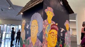 A small replica farmhouse is in the middle of a museum lobby. People look at it and its walls which are covered with murals of faces of the Nikkei farmers the installation honors. The faces are bright yellow and are against a dark night sky background with cranes flying. Vegetables and strawberries are seen throughout the murals too.