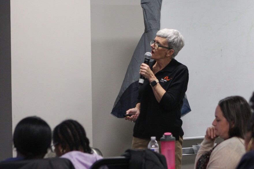 Julia Chalker speaks into a microphone in front of a group of disability advocates. She wears a long-sleeve black polo shirt with a small Arc of DFW Area logo on on the right side of her chest. She has a pixie-style hair cut with white and grey hair and wears glasses with a black frame. She is holding a clicker in one of her hands as she explains what is on the slide.