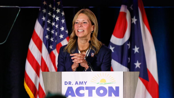 Dr. Amy Acton stands at a podium with a sign reading "Dr. Amy Acton for Governor." She is speaking in front of American and Ohio state flags.