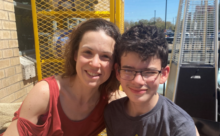 A woman smiles with her son who is wearing a Pokémon shirt. They are seated outside at a table in the sunshine with a glass of soda.