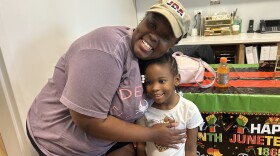 Sinquese Savage, a Black woman wearing a tan baseball cap and purple t-shirt, smiles at the camera while hugging her daughter, a little girl with her hair tied up with white beads at the ends of her braids. They stand in the entryway of a small daycare center that has a celebratory Juneteenth banner.
