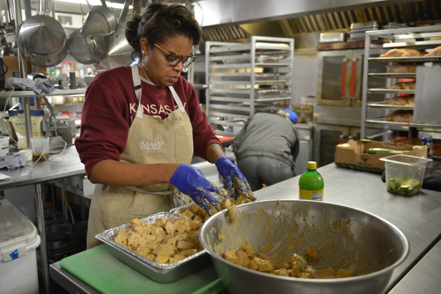 A woman wearing an apron works inside a large kitchen. She is wearing latex gloves and mixing a large bowl of moistened potatoes.