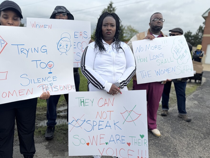 A group of protesters stand outside the women's huron valley correctional facility holding handmade signs. A woman in the center holds a sign that reads, "They can not speak So WE ARE THEIR VOICE!!!" Other signs read, "Trying TOO SILENCE WOMEN PRISONERS" with a drawing of a crying face, and "NO MORE!!! HOW LONG SHALL THE WOMEN Suffer!!"