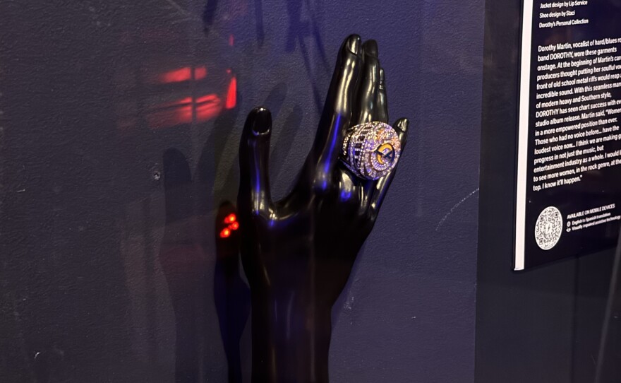 Displayed within museum glass, a black hand mannequin wears a spherical silver ring covered in white stones. In the center, gold lettering reads "LA."