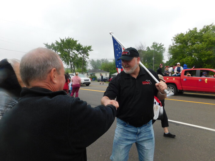 A white man wearing a black polo shirt and baseball cap and carrying an American flag shakes the hand of another white man along a parade route.