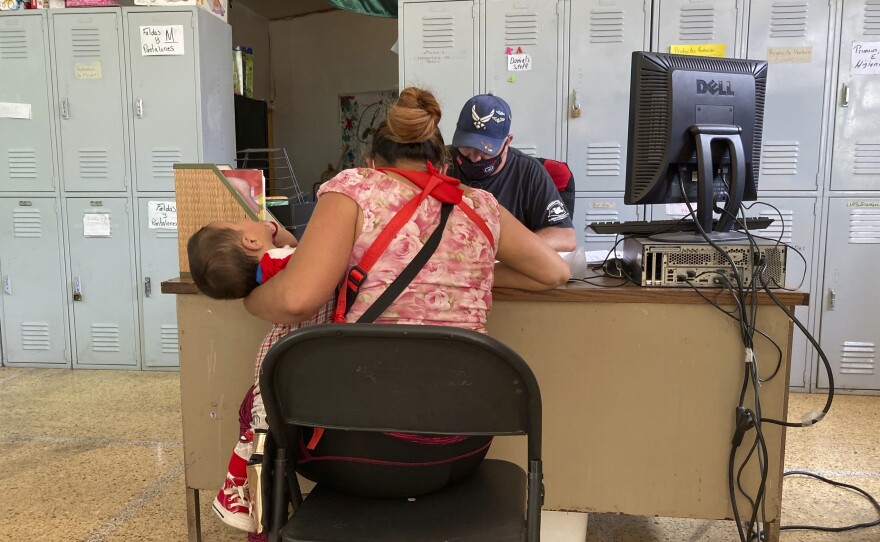 A woman sits at a desk with a young child on her lap. Behind the desk are lockers and boxes of items including diapers. 