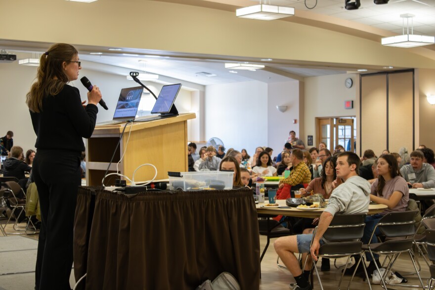 A woman speaks at a podium in front of a full room of people.