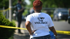 Outdoors street scene in daylight shows a Black woman from behind with her left hand resting on her hip and her right hand holding a box of tissue paper on her right hip. In background a police officer can be seen in the shadows with yellow police tape blocking a street.