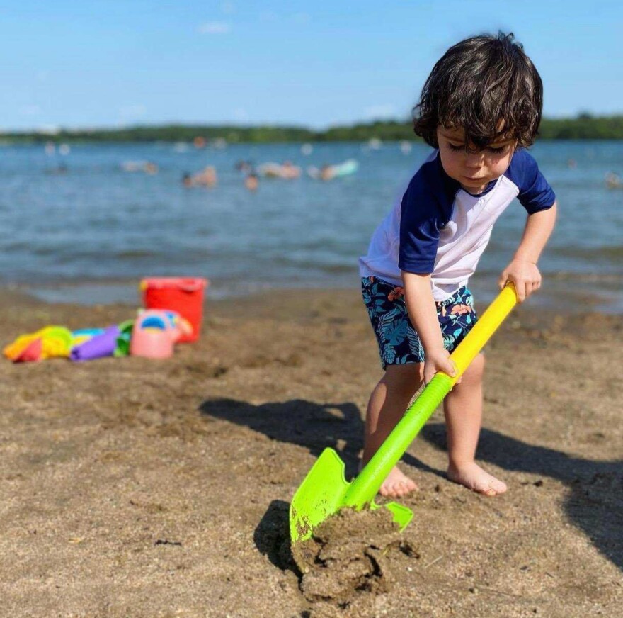 A toddler-age boy with curly brown hair shovels in the sand with a neon green plastic shovel.