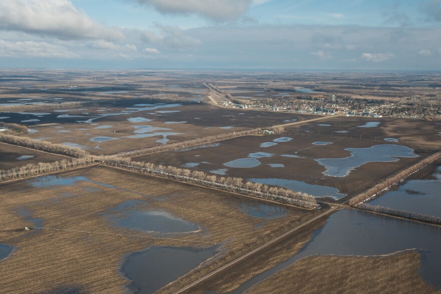 Floodwaters cover crop fields between roads lined with trees. A small town sits on the horizon.