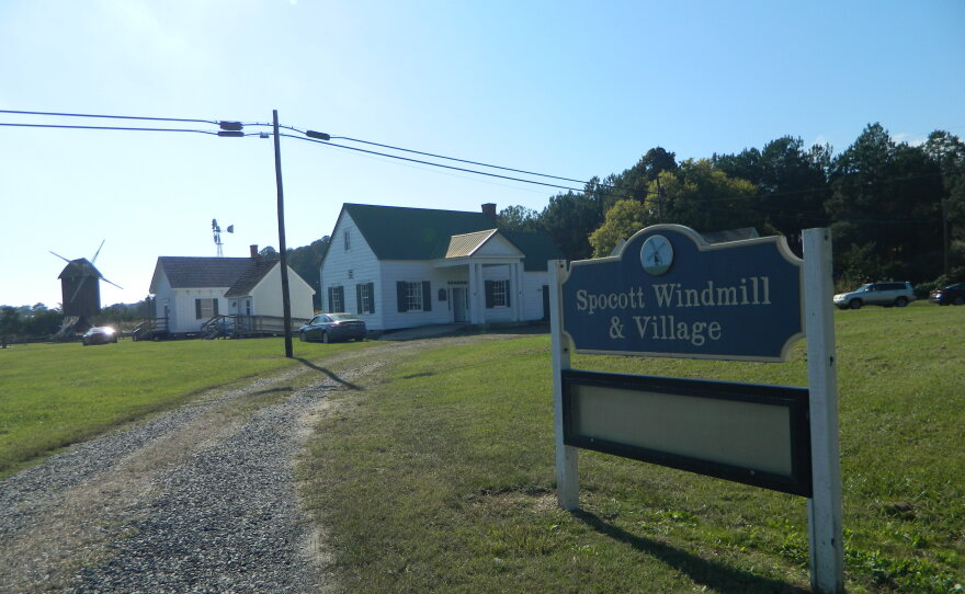 Entrance of the Spocott windmill and village. A large sign to the right of a gravel road welcomes visitors. In the background a couple simple buildings and the windmill at the farthest.