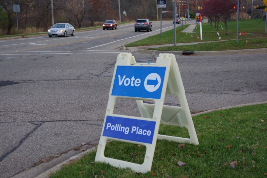 A blue and white sandwich board sign on the edge of an intersection with cars passing in the distance