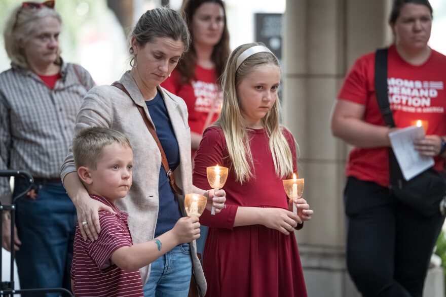 A photo of a woman with her arms around two children. All of them hold lit white candles and look solemn.
