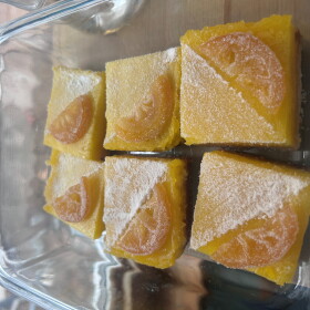 A clear pyrex plate of lemon bars with powdered sugar ana a lemon slice.