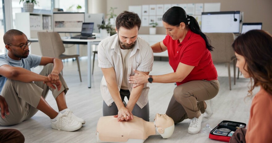 A woman coaches a man doing CPR chest compressions on a mannequin.