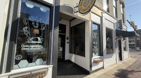 The front of Bee Joyful shop is depicted on the Kalamazoo Mall. The sidewalk in front of the store is red brick, and the storefront has two large glass windows with murals. One mural is of a cartoon bar of soap with a face and hands that is waving. Underneath it are the words “clean conscience”. The Bee joyful shop sign is a yellow circle with a bee hive in the middle.