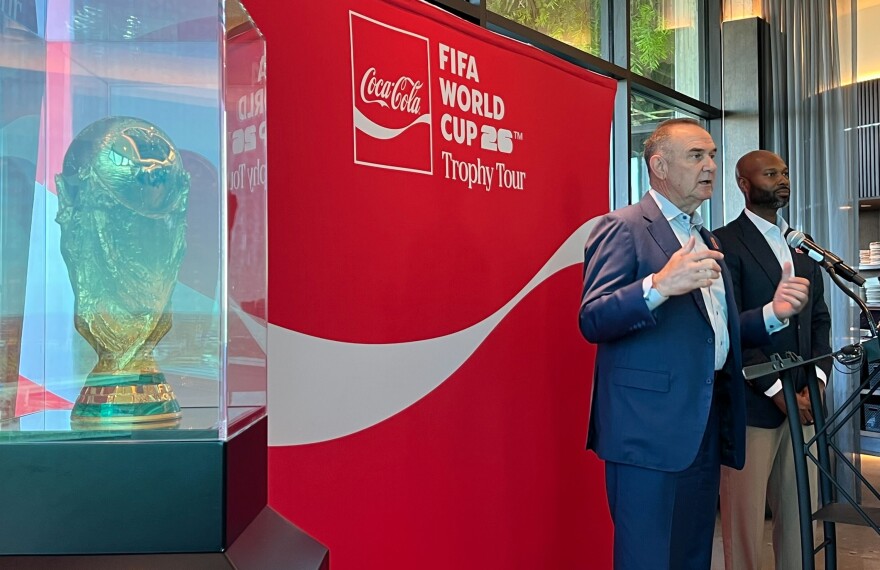 The World Cup trophy sits in a protective case as Missouri Gov. Mike Kehoe talks about the expected local impact from World Cup matches at Arrowhead Stadium. To Kehoe's left is Justin Bridgeman, CEO of Heartland Coca-Cola Bottling Company, which is sponsoring the trophy tour.