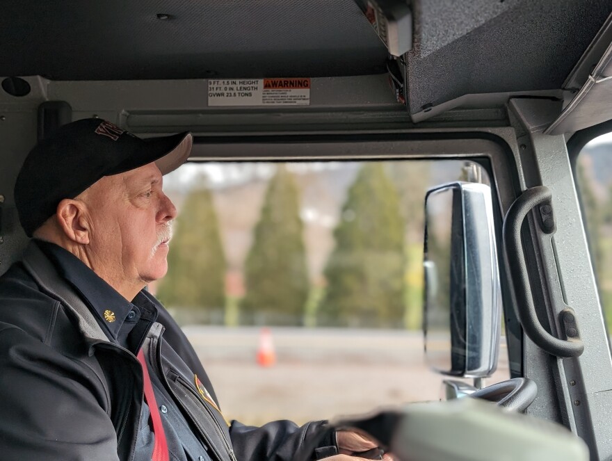 A man wearing a baseball cap sits in the drivers seat of a fire truck. He has a neutral expression as he drives the truck. The background is blurry but tall bushes and concrete are visible.