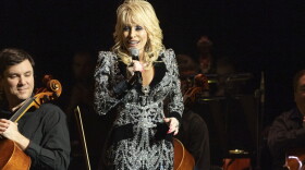 Dolly Parton stands on stage holding a microphone wearing a bedazzled outfit. 