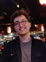 Headshot of Gustavo Sagrero. He is sitting in a restaurant while looking toward the camera and smiling.