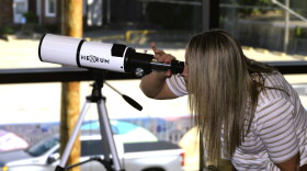 A woman's blonde straight hair drapes down, as she bends to peer through a white telescope, grasping it with her left hand. The instrument, aimed out a large glass window, has black lettering that reads Hexeum.