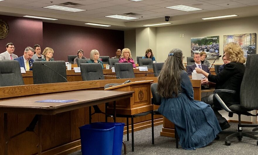 A blonde-haired woman sits before a committeee of legislators, speaking into a mircrophone. A woman in a long blue dress is sitting next to her.