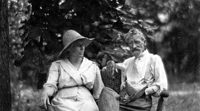 A black and white photograph of artist T. C. Steele and his wife Selma Neubacher Steele sitting outdoors on a wooden bench or chair. Selma, wearing a light-colored dress and sun hat, sits on the left, looking toward Steele. Steele sits on the right, wearing a light-colored shirt, suspenders, and boots, with one leg crossed over the other and his foot resting on the bench. They are seated in a grassy area with dense foliage behind them. 