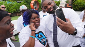 Black man in white dressshirt and tie  leans over to take a smartphone selfie with a shorter black woman in a white tshirt