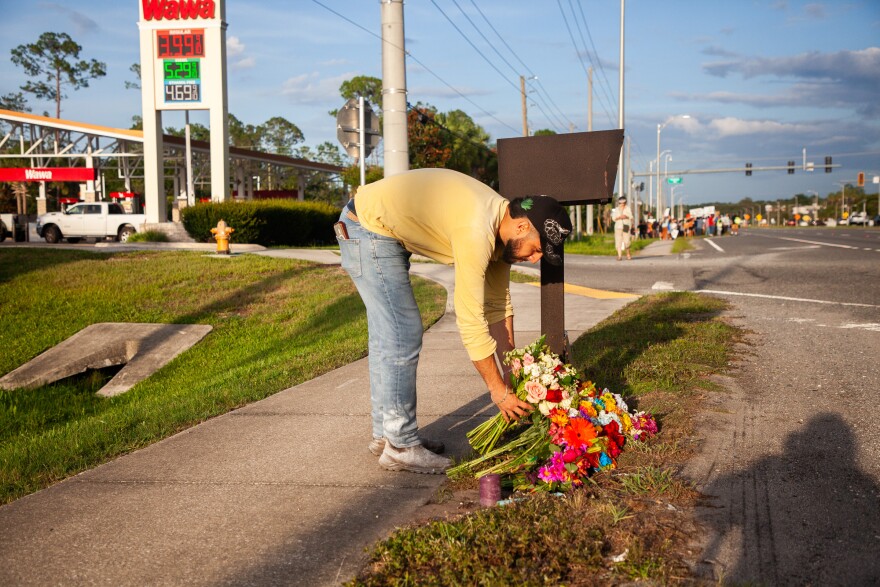 Man leans down to side of road to lay a bouquet of flowers down. A Wawa gas station is behind him 