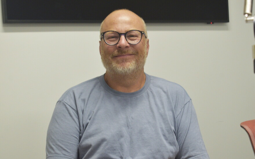 Man wearing glasses and a gray t-shirt seated in a radio studio with a monitor on the wall behind him.