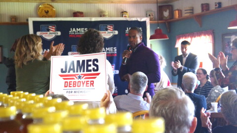 John James, a black man wearing a purple quarter-zip shirt, stands among a crowd of people who are clapping -- some are on their feet, and some are waving signs that say "James + Deboyer for Michigan". He is in a room with farmhouse-themed decorations, and there is a large banner behind him that reads "John James for Governor, Michigan". 