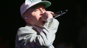 Man with a white baseball cap and brown sportcoat singing into a microphone